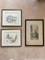 Three framed antique art prints on a carpet background, two with silver frames and one with a black frame, showing architectural and street scenes.
