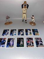 Three baseball figurines and ten baseball cards laid out flat on a white surface. Figurines are missing bats and have visible damage.