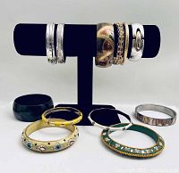 Photo of assortment of 10 bangle bracelets displayed on and around a black bracelet holder, showing variations in metal tones, enameling, and ornamental features.
