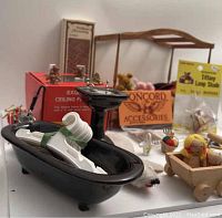 Photo of dollhouse miniature black bathtub with a white item inside, black pedestal sink, wooden bed frame, boxed ceiling fan light, Tiffany lamp shade packaging, and various miniature toys and accessories including teddy bears and a wooden toy cart with doll.