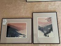 Pair of framed landscape prints with black frames and off-white mats displayed on carpet, showing misty valley mountains and coastal scene with soft pastel colors.