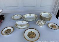Fine china pieces on a table, including plates, bowls, serving dishes with rose floral pattern and gold trim, alongside decorative embossed plates.