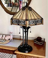 Full view of Tiffany style table lamp on wooden surface, showing hexagonal cream glass shade with black filigree accents and vintage bronze ornate base with pull chains.