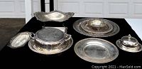 Top view of a collection of silver plated serving pieces including bowls, platters, and butter dishes arranged on a black cloth against white background.