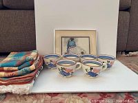 Six Stangl Pottery tea cups arranged in front of a framed art print with place mats folded to the side on a carpeted floor.
