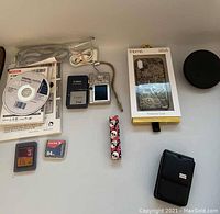 Canon camera with charger, instruction booklet, software CD, memory cards, small power pack, iPhone X/Xs protective case, and black circular case laid out on table.