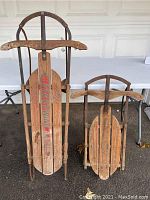 Photo showing two vintage wooden sleds with metal supports and runners, displaying rust and patina, stacked upright