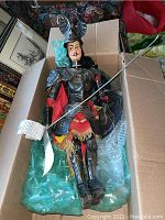 Full view of vintage knight marionette puppet lying in a box with protective bubble wrap, showing metal armor, red cape, sword, and shield with original hang tag.