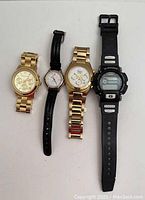 Four watches laid out flat showing entire fronts and straps including two gold-tone metal link watches, one black resin Casio G-Shock digital watch, and one ladies Wenger watch with leather strap.