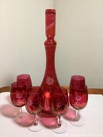 Cranberry glass decanter with stopper surrounded by six matching footed glasses on a table, showing the full set and color.