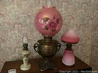 All three hurricane lamps arranged side by side (small white floral lamp, mid-sized Reform pink glass lamp, large Juno brass lamp with pink globe)