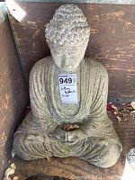Full frontal view of large concrete Buddha statue seated with closed eyes and draped robe, placed against wooden background, showing natural weathering.