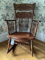 Front view showing carved wooden back panels with scroll and leaf patterns, turned spindle back and arm supports, seat with worn leather upholstery, and turned front legs.