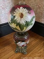 Front view of ornate hand painted duplex oil lamp showing large white flower with green leaves on top globe and matching floral on bottom globe with decorative metal base.