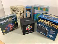 Photo showing all six items: three boxed new board games (On the Origin of Species, Subatomic, Periodic), Rubik's Touch Cube box, Our Amazing Home Planetarium box, and Jumbling Towers game in plastic container.