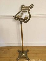 Full view of the vintage heavy brass music stand standing on a hardwood floor against a white wall. Shows entire structure including base, pole, and music rack shaped like a lyre.