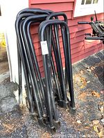 Ten black metal wood stacking brackets standing upright outdoors near a red wall, showing general wear.