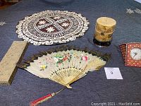 Overview of the antique hand-painted feather fan with bone reeds, decorative candle, and small clock displayed on a blue tablecloth with lace doily.