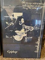 Black and white poster of John Lennon playing guitar with Epiphone logo and handwritten quote and signature in frame.