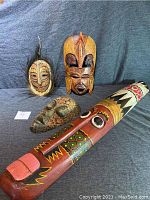 Four wood masks displayed on a gray backdrop showing distinct colors and carving styles, with one long carved cane in front