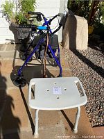 Blue 4-wheeled walker with padded armrests, hand brakes, front basket, and wooden cane beside white plastic bath stool with side handles