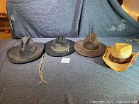 Four cowboy hats displayed side by side on a blue-gray fabric background, including three felt hats in black and brown tones and one straw hat with decorative band.