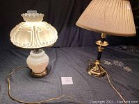 Pair of lamps shown on a table with dark cloth backdrop; milk glass lamp with scalloped top and brass base on left, brass candlestick lamp with beige pleated shade on right.