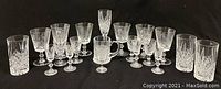 Seventeen assorted crystal beverage glasses on black background, including various stemmed glasses and tumblers with cut patterns.