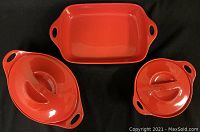 Three CorningWare red stoneware pieces displayed together on black background: small circular pot with lid, larger oval pot with lid, and rectangular baker.