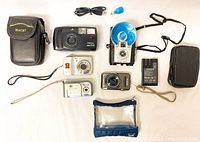 Seven assorted cameras along with accessories including battery charger and carrying cases arranged on a white background