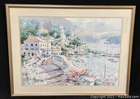 Framed watercolor-style print depicting a coastal village with boats, bridge, buildings, flowers, and harbor scene. Signed by artist in the bottom right.