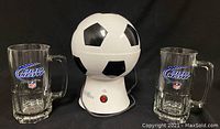 Front view of the soccer ball popcorn machine centered between two clear Bud Light NFL pint glass mugs, all items on black surface.