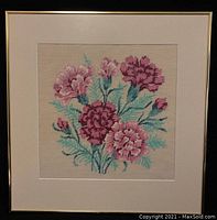 Front view of the needlepoint tapestry showing pink flowers and green leaves, framed in a thin metal frame with off-white matting.