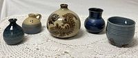 Front view of 5 pottery pieces arranged side by side on a crocheted tablecloth, showing various shapes, colors, and decorative elements