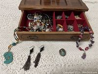 Open wooden jewelry box showing many compartments filled with assorted costume jewelry including necklaces, earrings, rings, and bracelets on white crocheted surface.