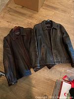 Two men's Eddie Bauer leather jackets, brown and black, laid on wooden floor showing front and collar details