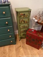View showing both the olive green five-drawer cabinet with detailed painted panels and the red two-drawer cabinet with floral motifs on the floor beside it.