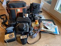 Grouping of cameras, flashes, binoculars, camera case, instruction books and other camera accessories shown on wooden surface
