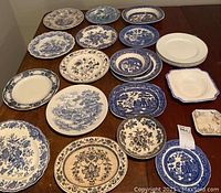 Top view of collection of 18 china plates featuring blue and white transferware patterns with floral and scenic motifs, arranged on wood table.