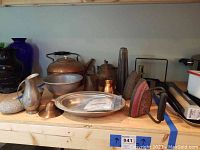 Full view of all items on wooden shelf including the copper kettle, vintage irons, metal candle maker, pots, bowl, jug, and rock.