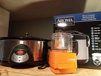 Photo showing Aroma rice cooker in its box, Cuisinart mini prep processor, and Crockpot slow cooker on wooden shelf.
