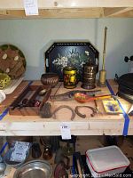 Wooden shelf with multiple brass decorative items, three horseshoes, wooden ladle with painted design, metal jars, and three pipes displayed, some items look aged and dusty.