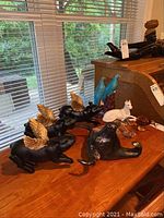 Photo showing 3 black flying pigs with gold wings, 2 blue birds on a branch, white dog, black bull head, and small other figurines arranged on a wooden table in front of window.