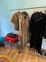 Two full-length coats and one short mink stole on a garment rack. One coat is light brown fox fur, the other is a dark mink with plush collar and cuffs.