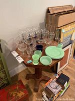 Assortment of clear glass stemware and green glass dishes arranged on octagonal wooden table near wall with various other household items nearby.