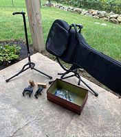 Two adjustable FretRest guitar stands, one Access soft guitar carry case with backpack straps, and two Hercules wall holders on wooden mounts arranged outdoors on concrete next to grass and wood post.
