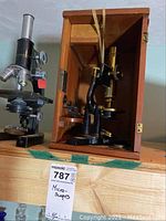 Two microscopes shown, one in wooden case with strap, missing door panel, and one black microscope with silver knobs.