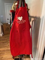Red apron with 'I ❤️ my Vita-Mix' white print hanging with other aprons and bags in background