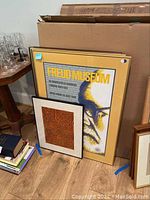 Two framed pieces leaning against cardboard boxes. The larger is a Freud Museum poster with yellow border and visible crack in the glass, smaller abstract framed artwork with orange geometric pattern in front.
