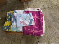 Four quilts folded on a chair showing variety of colors including bright yellow with X pattern, blue and white patchwork, light floral/pink pattern, and Iranian magenta quilt.